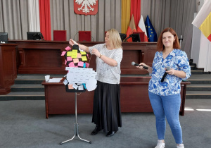 Prowadzące panie stoją przy stojaku z karteczkami